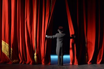 Image depicting rear view of lady in top hat and tails as she peers through parted red curtains to the stage and seats beyond.