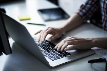 A person is typing on a laptop, their hands are visible, and their other hand is on the table. There is a notebook and a pen on the table.