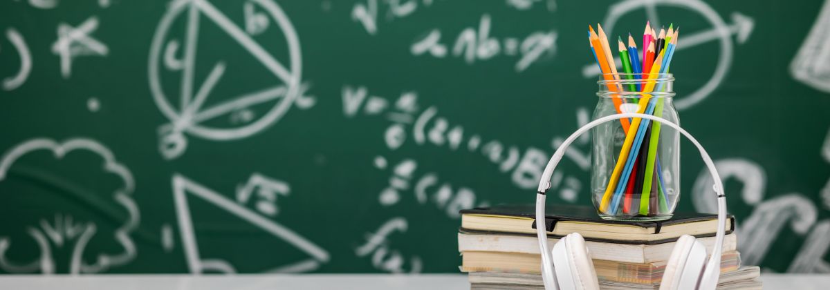 Headphones on a stack of books with a jar of colored pencils, in front of a chalkboard with math formulas.